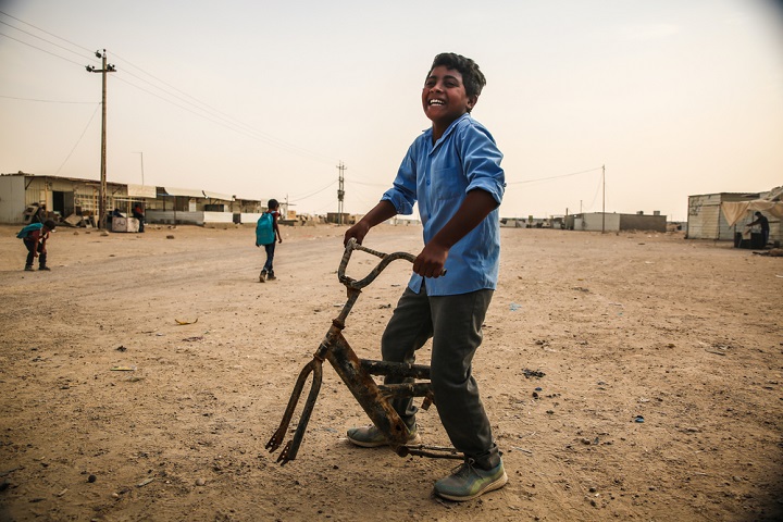 荒野にテントとコンテナが点在するアムリヤット・ファルージャ避難民キャンプで、壊れた自転車に乗って遊ぶ少年　© Mohammad Ghannam/MSF