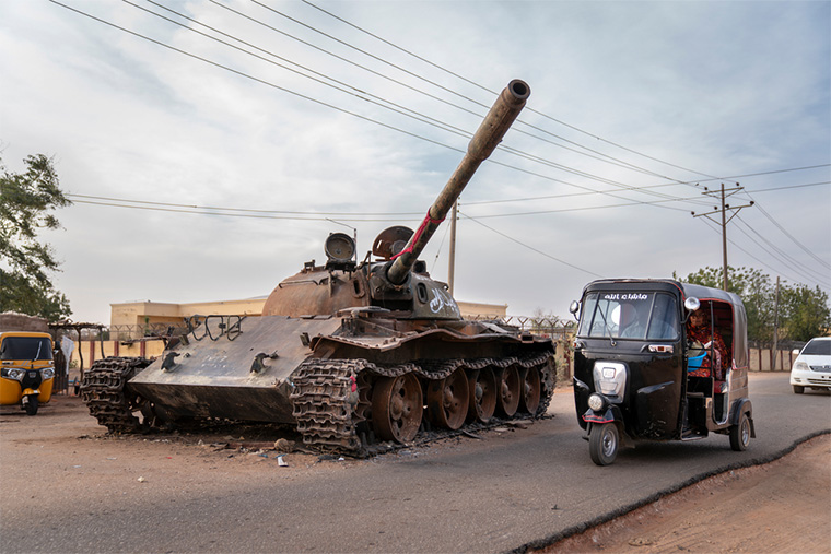 スーダン：破壊されたスーダン軍の戦車と、その横を走るリキシャタクシー。2023年、西ダルフール州のジェネイナで起きた激しい戦闘の名残だ＝2024年2月20日　Ⓒ  Diana Zeyneb Alhindawi