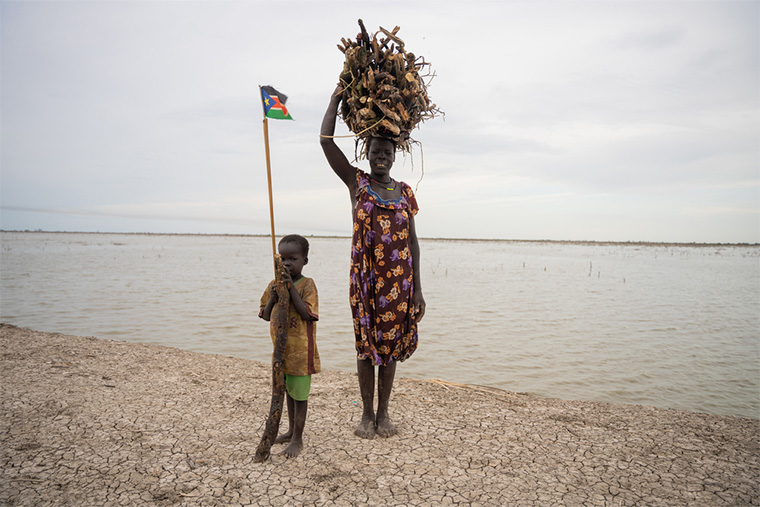 南スーダン：ユニティー州ベンティウの国内避難民キャンプ。洪水で流された木材を運ぶローダ・ネファザル・ムノットさんと、どこへ行くにも母親の後をついていく息子＝2024年6月21日　Ⓒ Isaac Buay/MSF