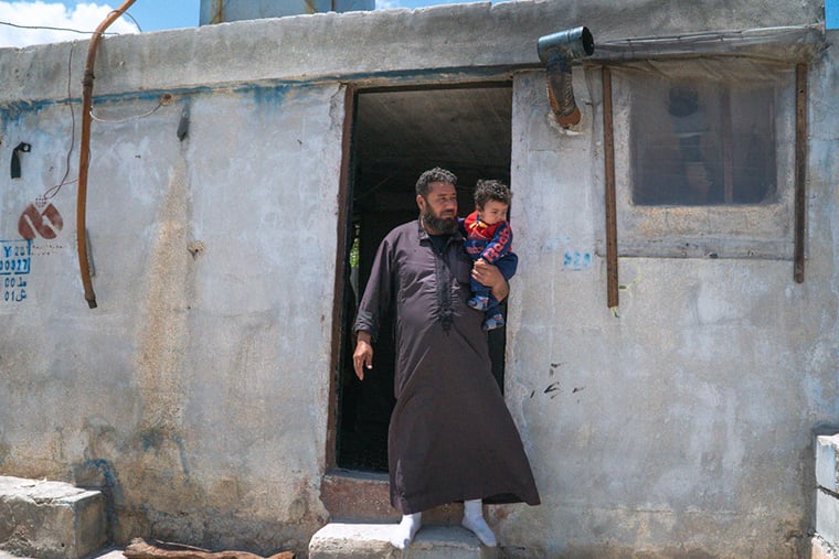 シリア／イドリブ県：アル・クウェイティ難民キャンプで避難所の外に立つワリードさんと末の息子。2024年12月初めのアサド政権崩壊以来、数千の避難民家族が帰還を試みてきたが、実現していない。数週間前に村へ戻ったワリードさん一家を待っていたのは、空っぽの廃墟と旧政権が残した地雷の残骸だった＝2025年5月　© Ahmad Amer/MSF