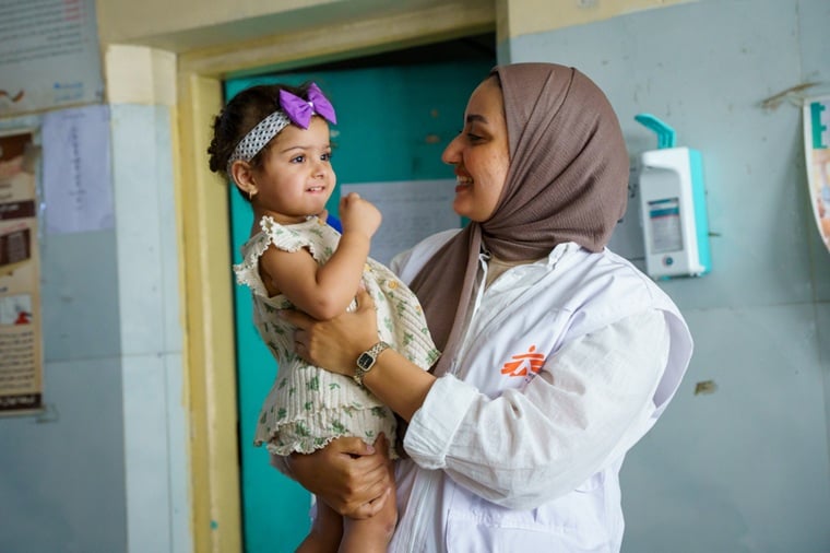 イラク／バグダッド ：サドルシティにあるアル・マフディ診療所で、結核の経過観察を受けているザフラちゃんを抱くMSFのヘルスプロモーター。MSFは地域の人びとに向けて、結核に関する啓発活動とスクリーニング検査を行っている＝2025年9月　© Deniz Fahmi/MSF