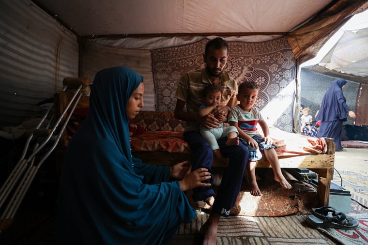 パレスチナ／ガザ地区：9カ月のモハメドちゃんと2歳半のイブラヒムちゃんを抱くバセルさん。その傍らで妻が、切断された脚の幻肢痛を和らげるためにマッサージをしている。バセルさんは数カ月前、配給所で支援物資を受け取ろうとした際に銃弾を受け、脚を失った。家族はラファの自宅を逃れ、ハンユニスのマワシ地区にある仮設テントで暮らしている＝2025年10月　© Motasem Abu Aser/MSF