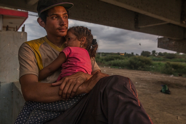 ペルーにあるトゥンベス川のほとりで娘を抱くベネズエラ人移民。ここで移民たちは休息をとり、日差しや雨をしのいでいる＝2022年　Ⓒ MAX CABELLO ORCASITAS