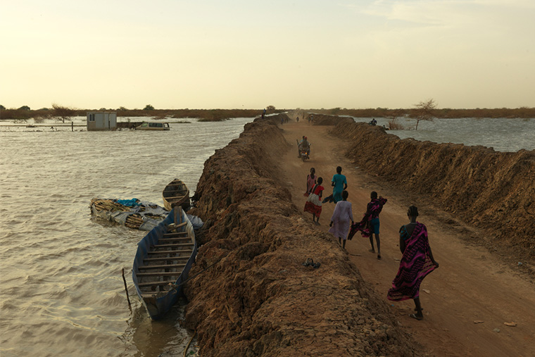 約12万人が暮らす南スーダンのベンティウ・キャンプで、堤防の間を歩く洪水避難民たち。この堤防がキャンプ全体の浸水を防いでいる＝2022年　Ⓒ Peter Caton