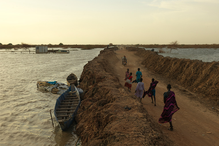 約12万人が暮らす南スーダンのベンティウ・キャンプで、堤防の間を歩く洪水避難民たち。この堤防がキャンプ全体の浸水を防いでいる=2022年 Ⓒ Peter Caton